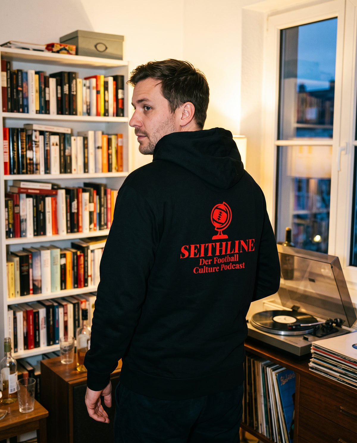 Fan im schwarzen Seithline-Hoodie mit Rückenaufdruck vor Bücherregal und Plattenspieler
